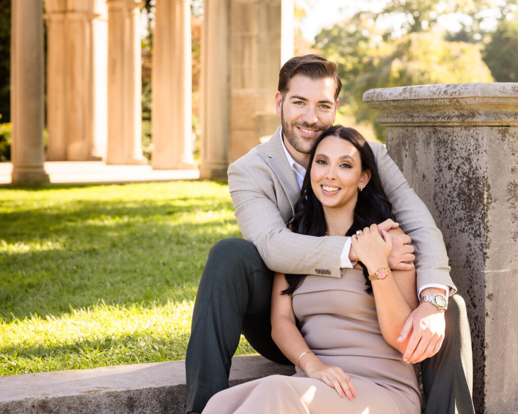 Romantic engagement photo of a couple sitting together near the elegant stone columns of Coe Hall at Planting Fields Arboretum in Oyster Bay, New York, bathed in warm afternoon sunlight.