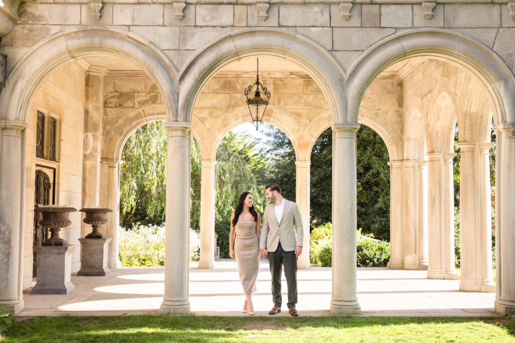 Elegant engagement photo of a couple walking hand in hand beneath the stone arches of Coe Hall at Planting Fields Arboretum, showcasing timeless Long Island estate architecture.