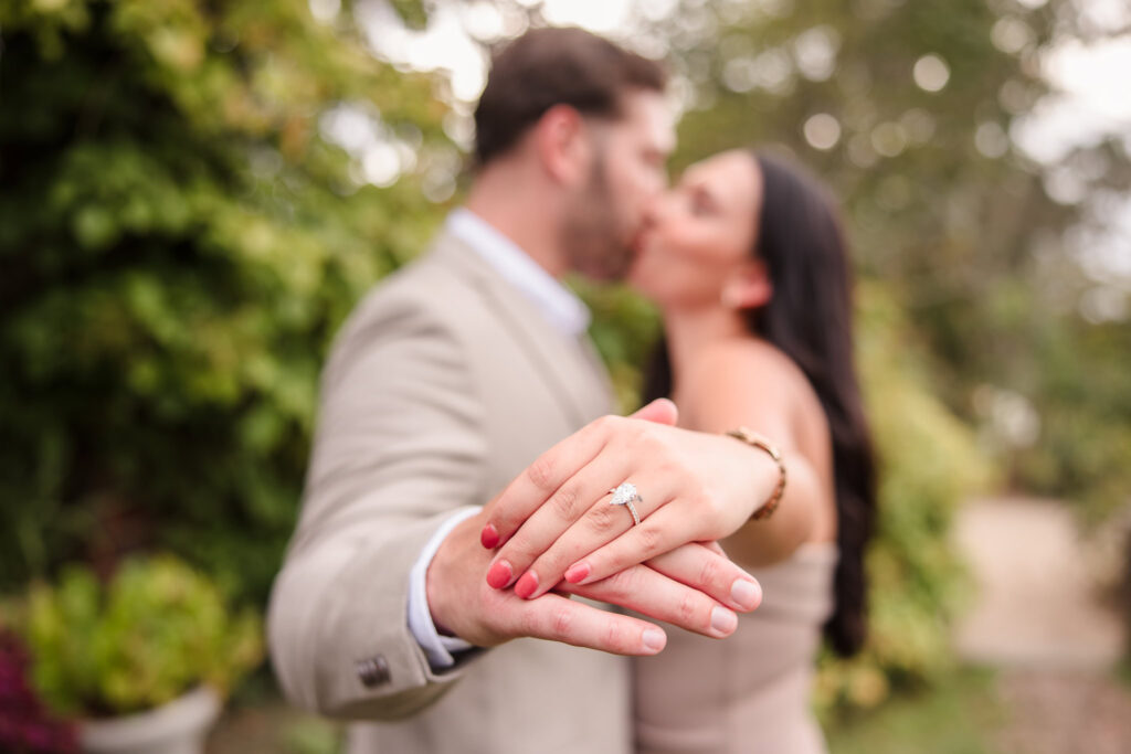 Close-up engagement photo highlighting a diamond ring as the couple kisses in the gardens of Planting Fields Arboretum, capturing romance and timeless elegance.