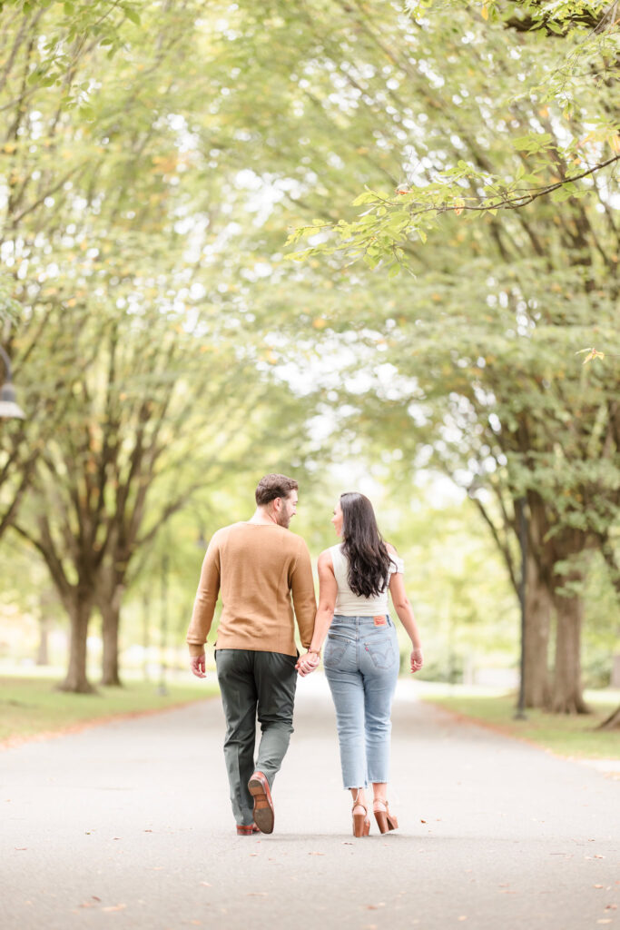 Engagement photo of a couple walking hand in hand down a tree-lined path at Planting Fields Arboretum, surrounded by soft greenery and romantic light.