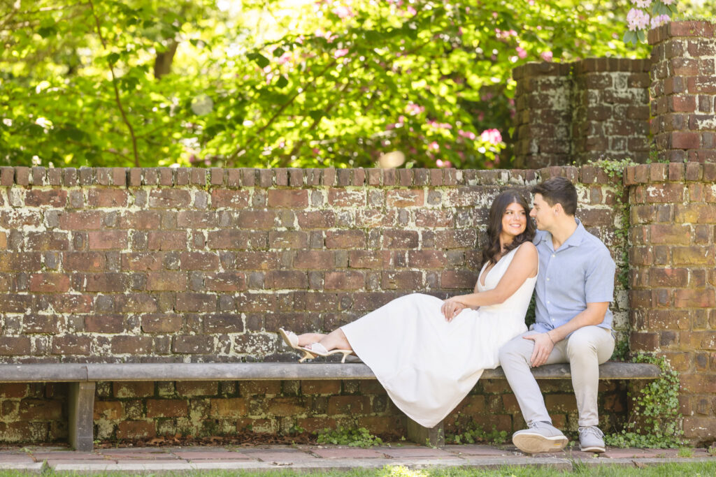 Engagement photo of a couple sitting on a stone bench by the brick garden wall at Planting Fields Arboretum, surrounded by lush greenery and soft sunlight.