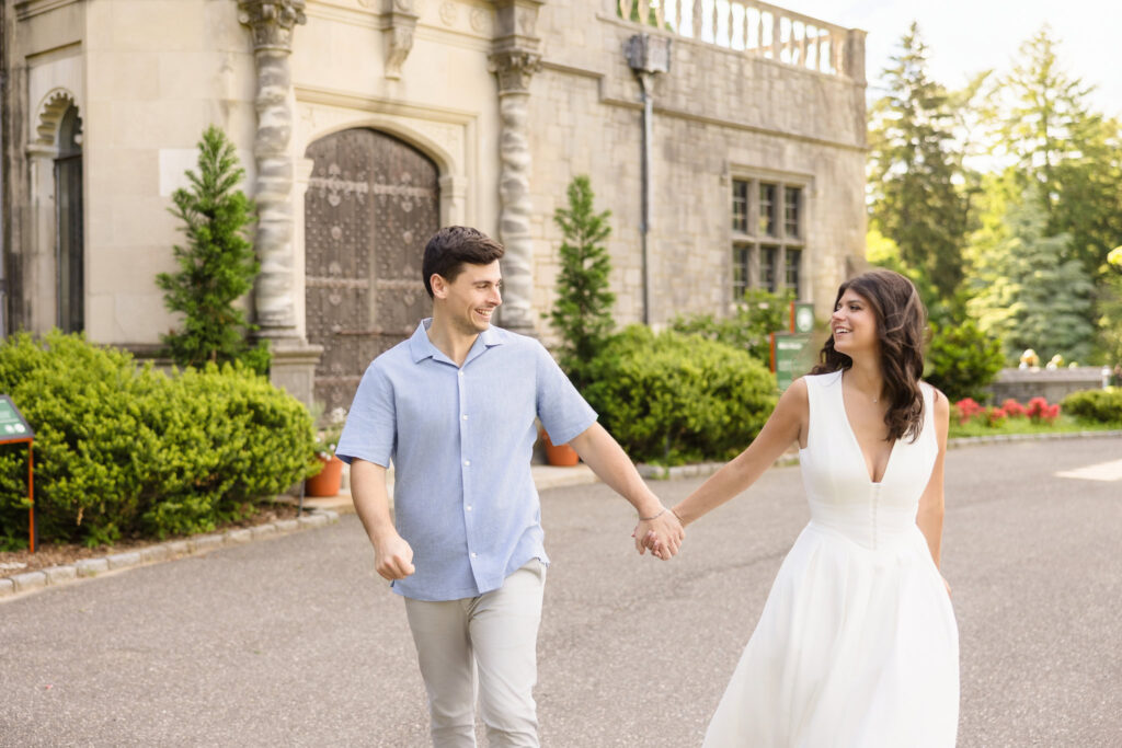 Joyful engagement photo of a couple walking hand in hand outside Coe Hall at Planting Fields Arboretum, showcasing the mansion’s elegant stone architecture and garden surroundings.