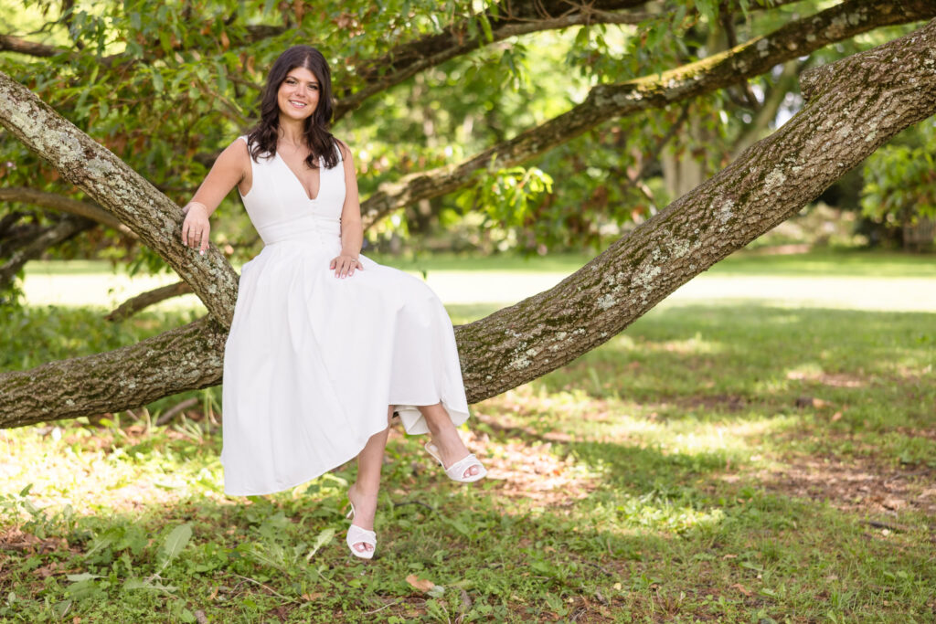 Bride-to-be sitting gracefully on a tree branch during her engagement session, surrounded by lush greenery and natural sunlight.