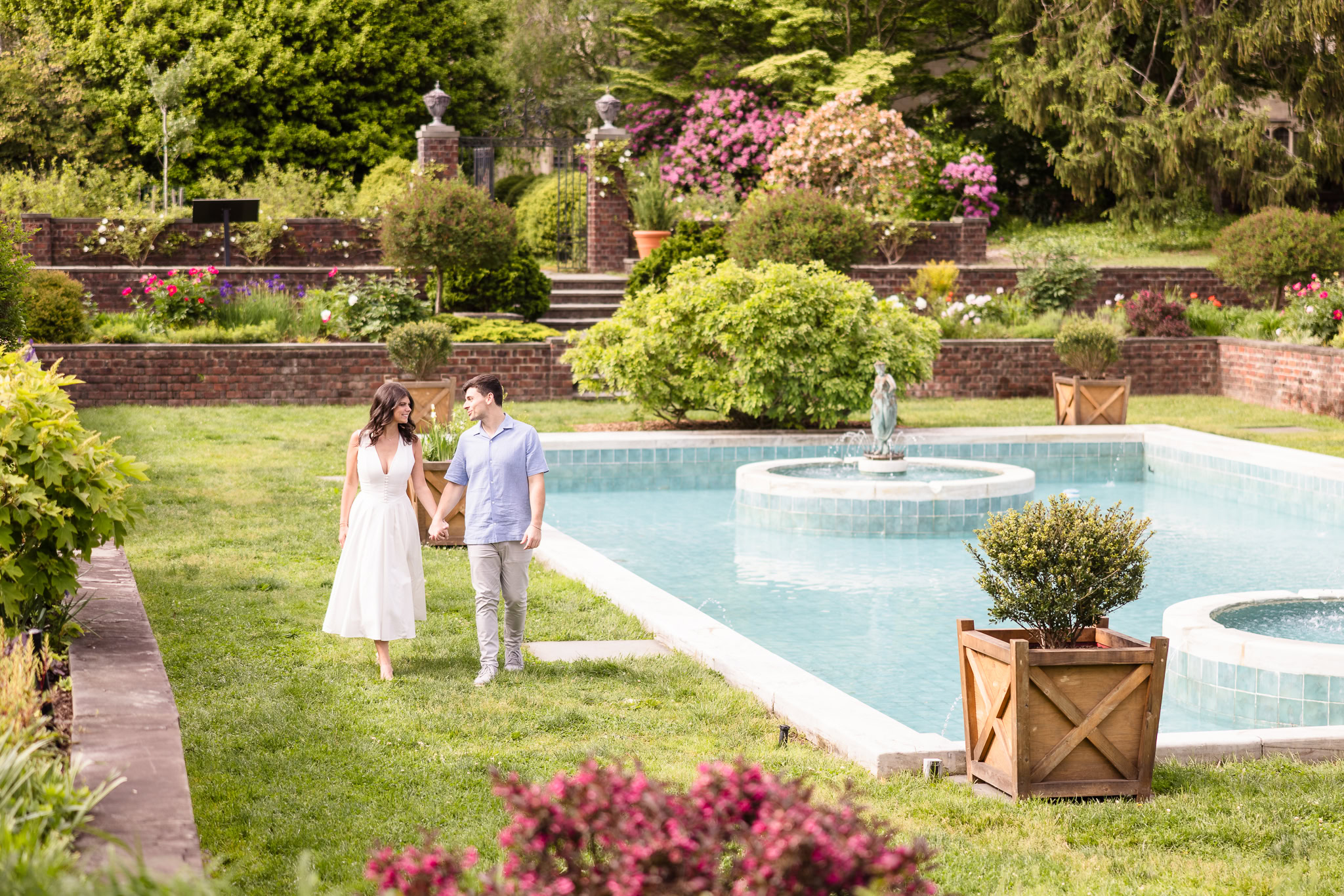 Engagement photo of a couple walking hand in hand beside the elegant reflecting pool in the Italian Garden at Planting Fields Arboretum, surrounded by blooming flowers and greenery.