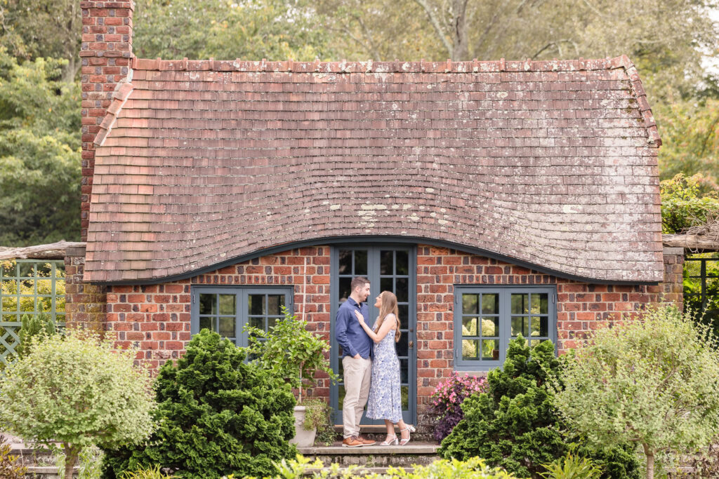 Charming engagement photo of a couple standing in front of the storybook brick cottage at Planting Fields Arboretum, surrounded by lush greenery and romantic garden details.