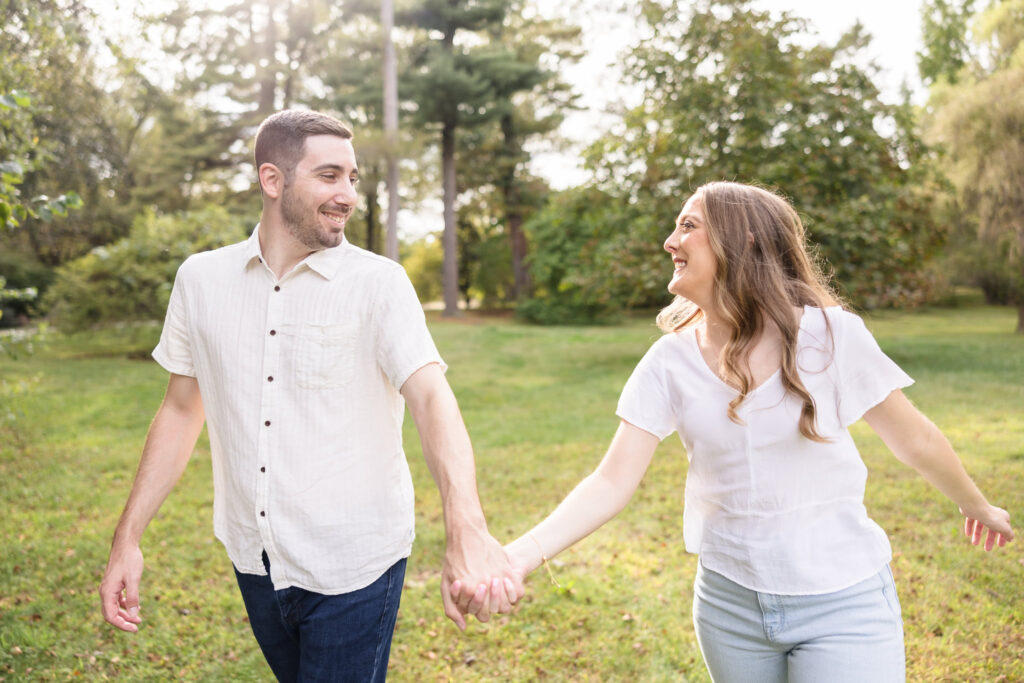 Joyful engagement photo of a couple holding hands and walking through the sunlit gardens at Planting Fields Arboretum during golden hour.