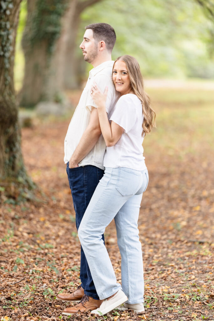 Bride-to-be embracing her fiancé from behind during their fall engagement session at Planting Fields Arboretum, surrounded by golden leaves and soft natural light.