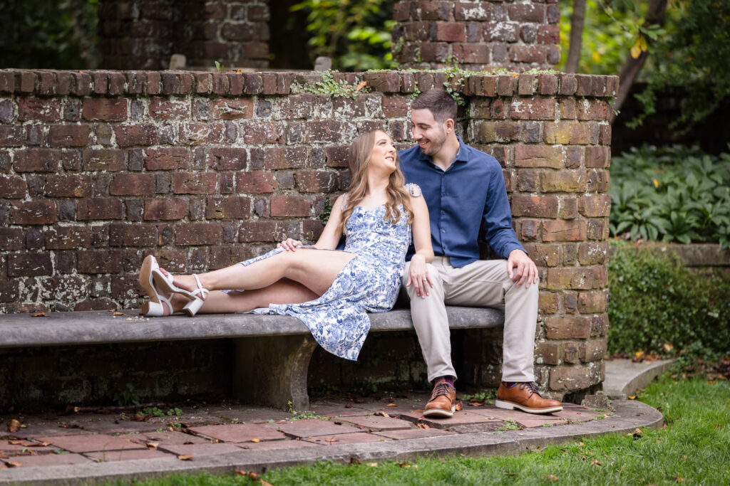 Couple sitting together on a stone bench against a rustic brick wall during their romantic fall engagement session at Planting Fields Arboretum in Oyster Bay, New York.