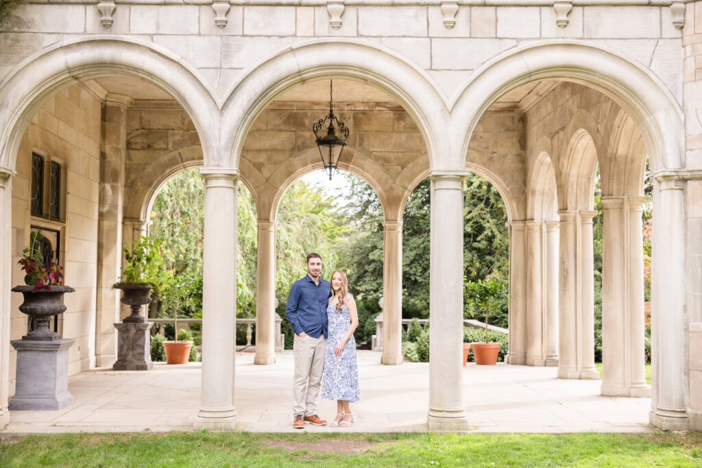 Engaged couple standing beneath the grand stone arches of Coe Hall at Planting Fields Arboretum during their elegant fall engagement session in Oyster Bay, New York.
