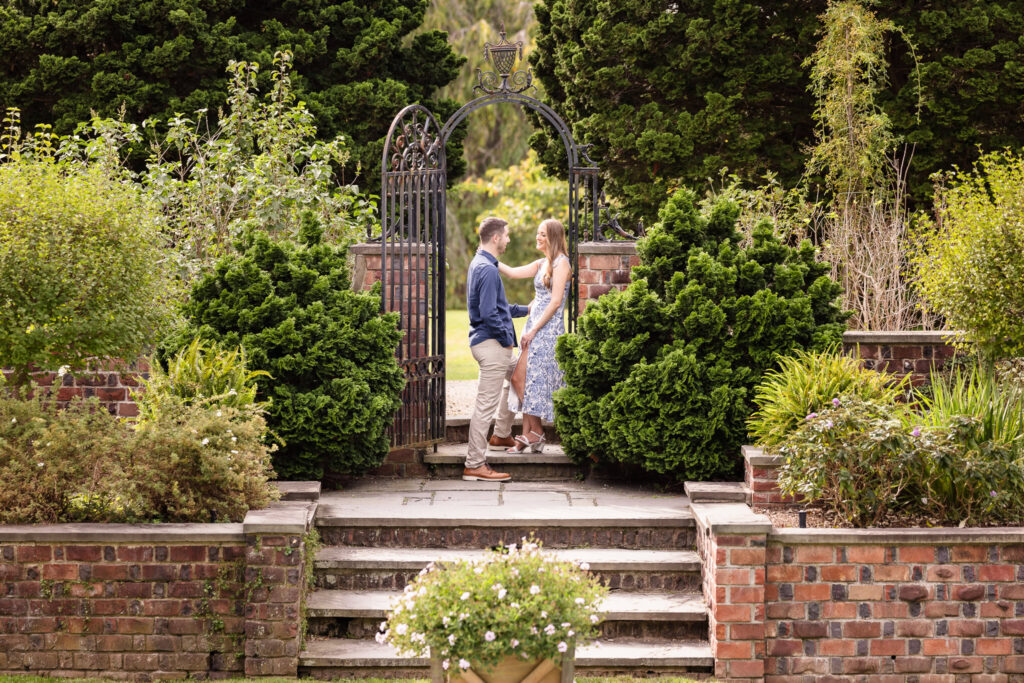 Couple shares a romantic moment by the wrought-iron gate surrounded by lush fall greenery at the Italian Gardens in Planting Fields Arboretum, Oyster Bay.