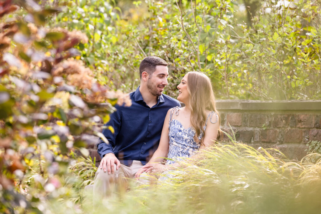 Couple sits together surrounded by golden fall foliage at the Italian Gardens during their romantic engagement session at Planting Fields Arboretum in Oyster Bay.