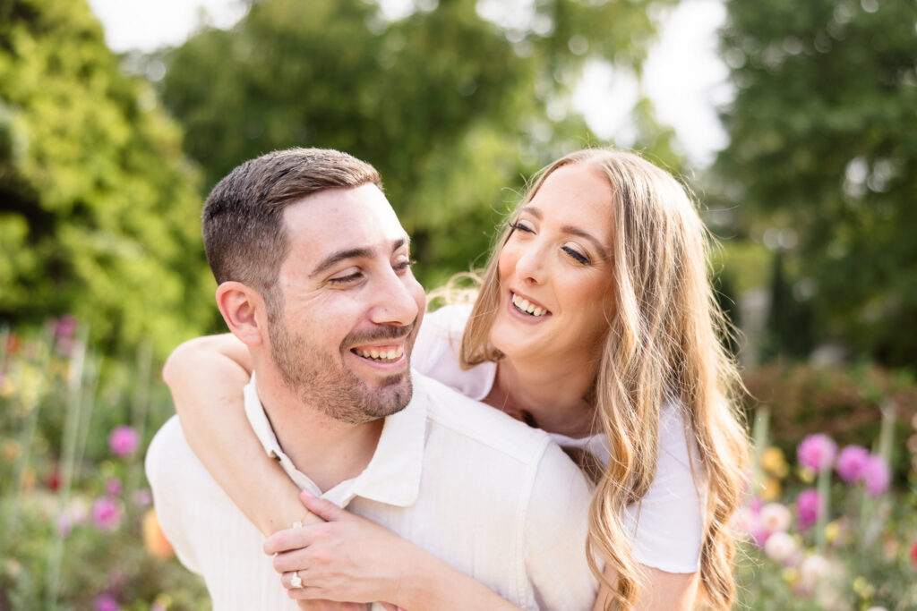 Couple shares a playful piggyback moment surrounded by colorful fall blooms during their engagement session at Planting Fields Arboretum in Oyster Bay, New York.
