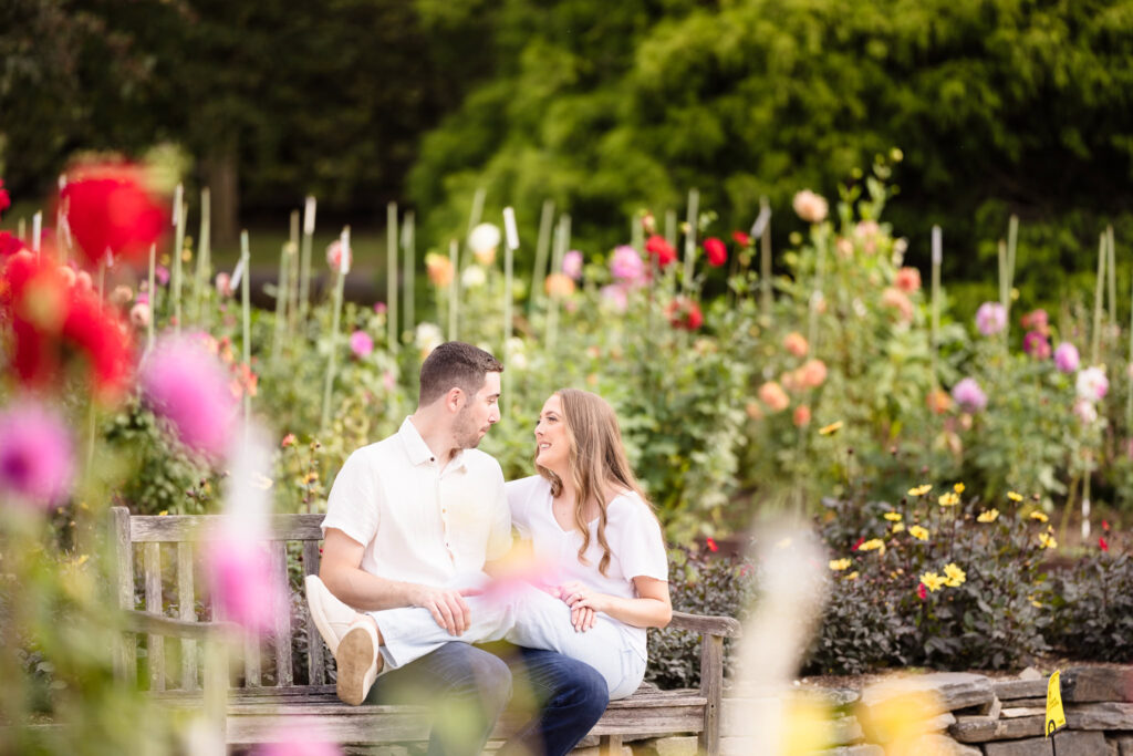 Engaged couple sitting together on a wooden bench surrounded by colorful fall flowers during their Planting Fields Arboretum engagement session in Oyster Bay, New York.