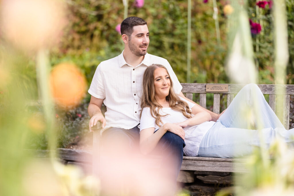 Couple relaxes together on a garden bench surrounded by soft fall florals during their engagement session in Oyster Bay, Long Island.