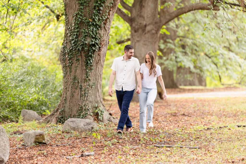 Engaged couple walking hand in hand beneath tall trees surrounded by early fall foliage during their Planting Fields Arboretum engagement session in Oyster Bay, New York.