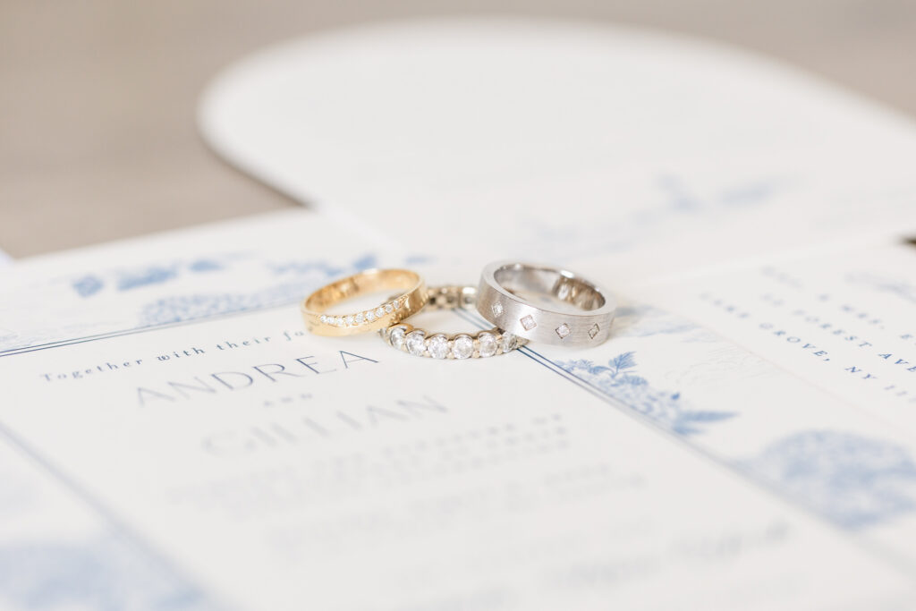 Close-up of gold and silver diamond wedding bands resting on Andrea and Gillian’s Snapper Inn wedding invitation in Oakdale, New York.