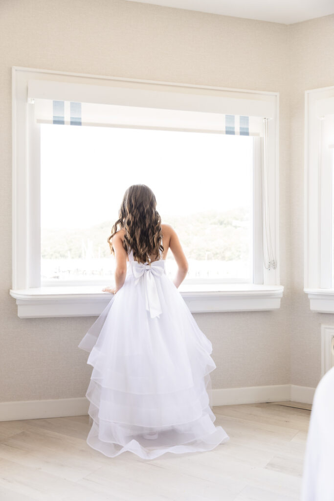 Flower girl in a tiered white dress with a bow looking out a bright window before Andrea and Gillian’s Snapper Inn wedding in Oakdale, Long Island.
