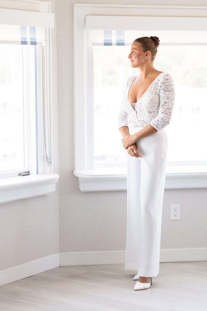 Bride standing by a bright window in a lace top and white jumpsuit, getting ready for her elegant waterfront wedding at The Snapper Inn in Oakdale, Long Island.