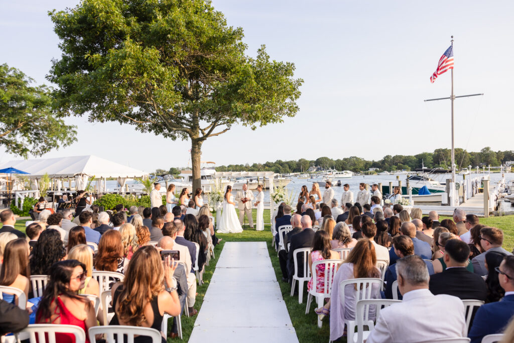 Outdoor waterfront wedding ceremony at The Snapper Inn in Oakdale, Long Island, with guests seated along the Connetquot River on a sunny summer day.