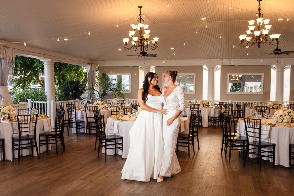 Newlywed brides sharing a joyful moment inside The Snapper Inn’s elegant reception pavilion in Oakdale, Long Island, surrounded by white floral centerpieces and candlelit tables.