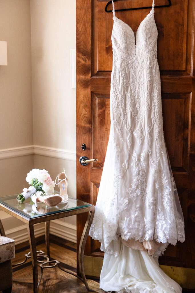 Elegant lace wedding dress hanging on a wooden door with bridal shoes and white bouquet nearby before an elegant summer wedding at Larkfield Manor.