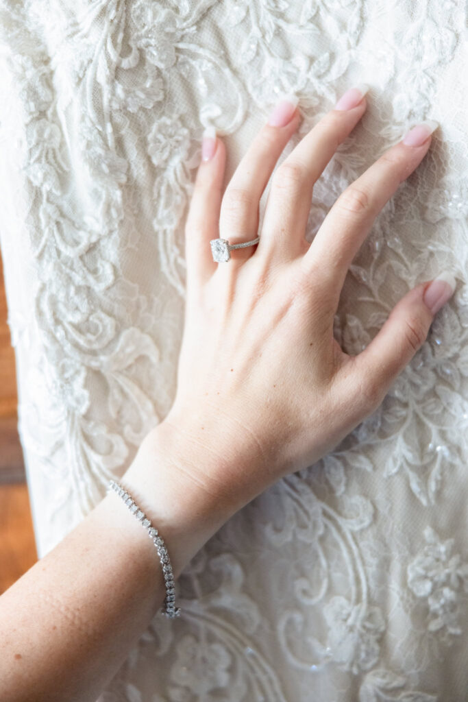 Close-up of bride’s diamond engagement ring and bracelet resting on lace wedding gown before elegant summer wedding at Larkfield Manor.