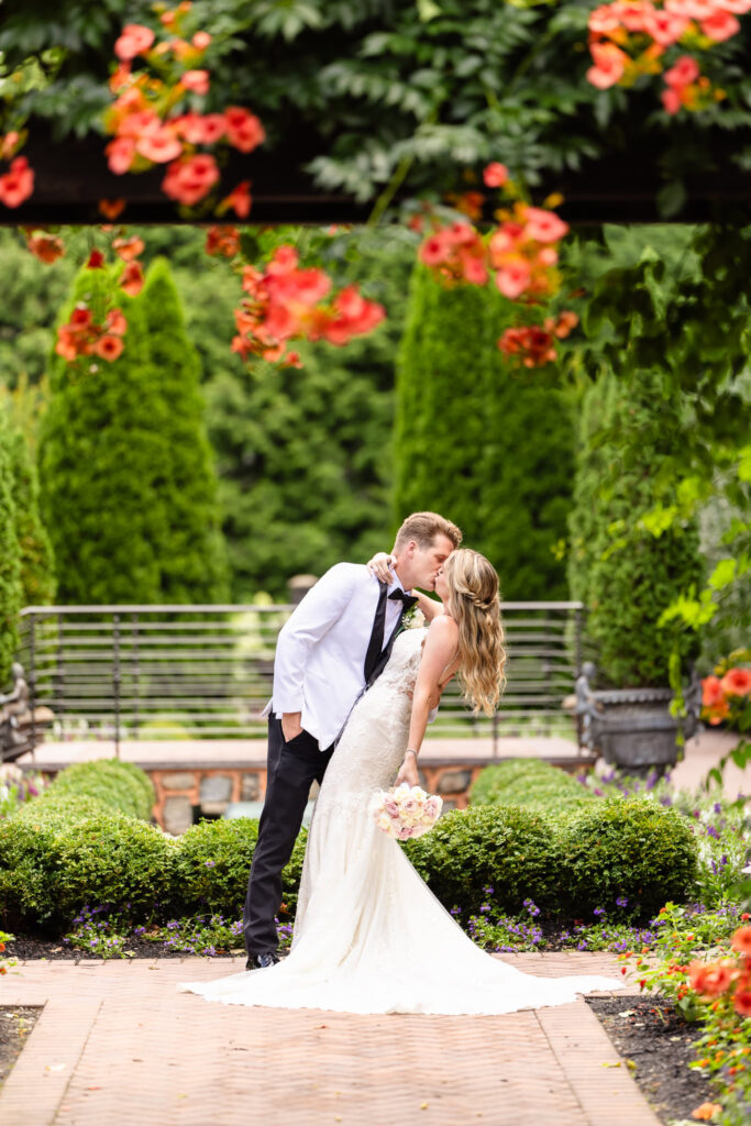 Bride and groom share a romantic kiss surrounded by lush greenery and vibrant flowers in the garden at Larkfield Manor during their elegant summer wedding.