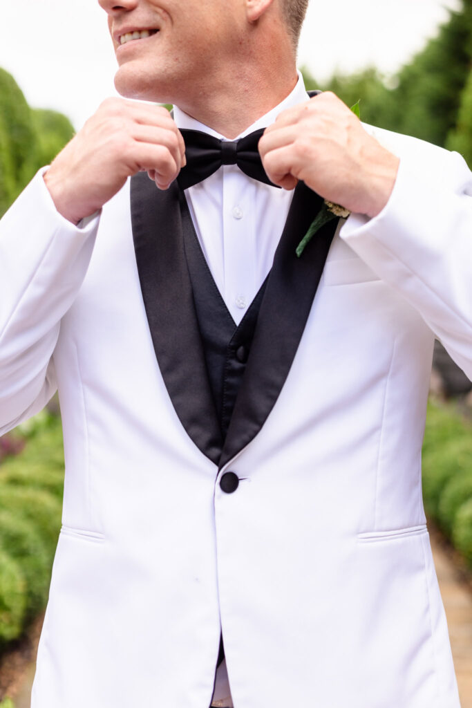 Groom adjusts his black bow tie while wearing a classic white tuxedo jacket during his elegant summer wedding at Larkfield Manor.