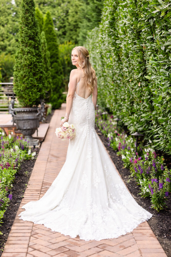 Bride in lace Martina Liana gown holding pastel rose bouquet and smiling over her shoulder in the lush garden at Larkfield Manor.