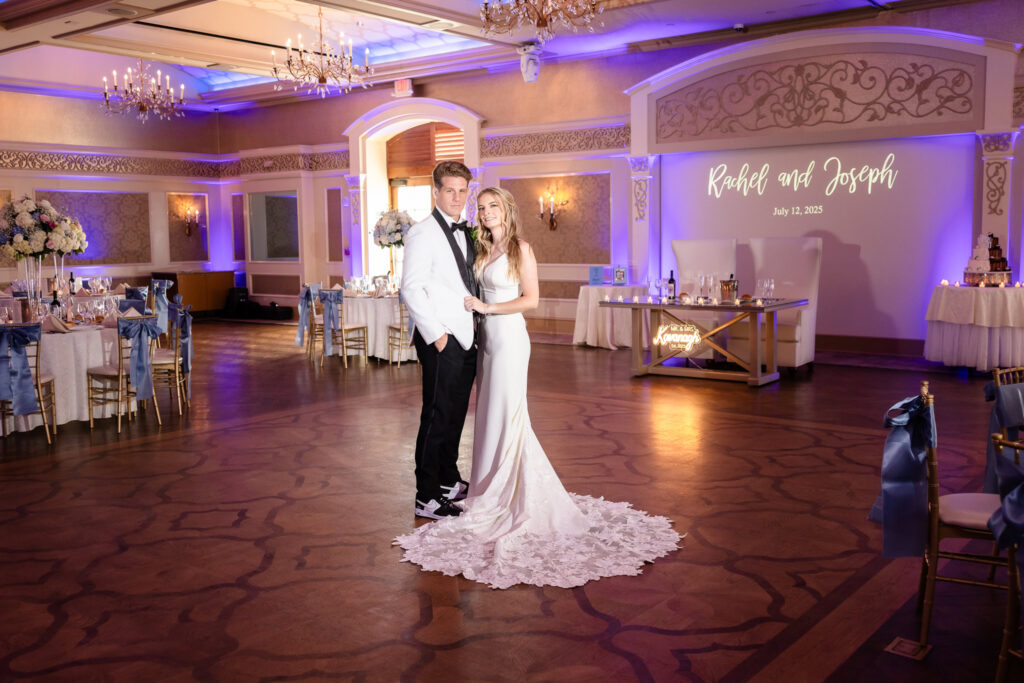 Bride and groom posing in the elegant Larkfield Manor ballroom with personalized monogram lighting, blue decor, and floral centerpieces for their summer wedding reception.