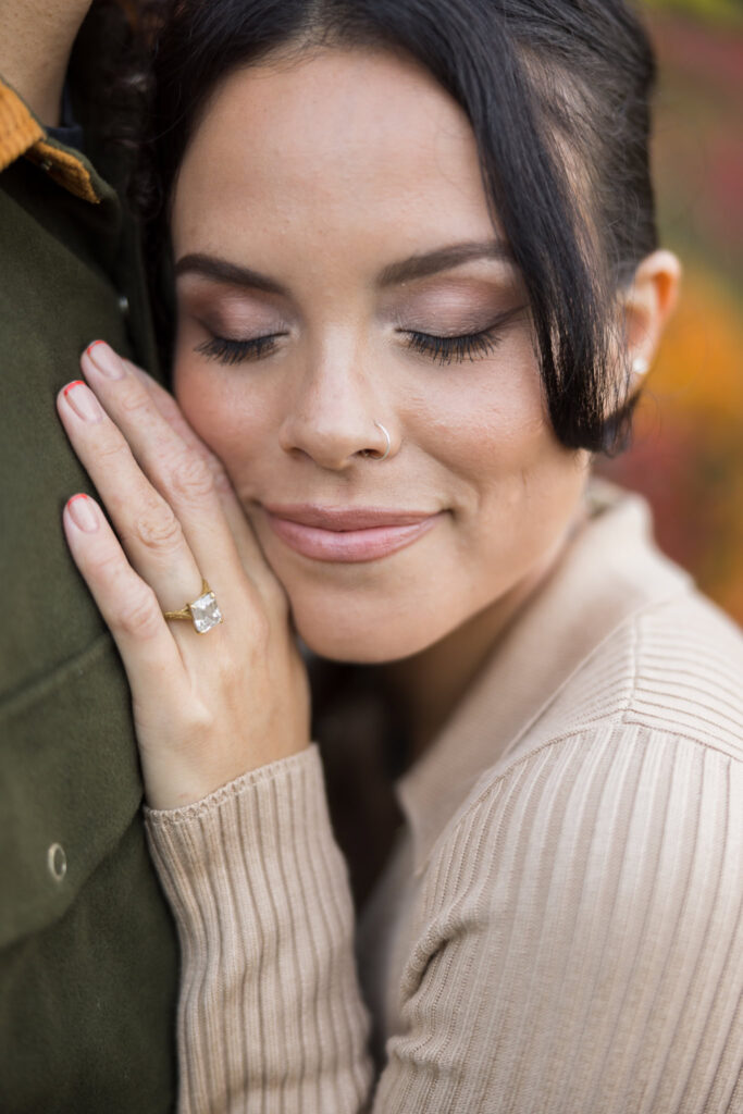 Close-up of bride-to-be resting her cheek on fiancé’s shoulder, showcasing her engagement ring during a fall session