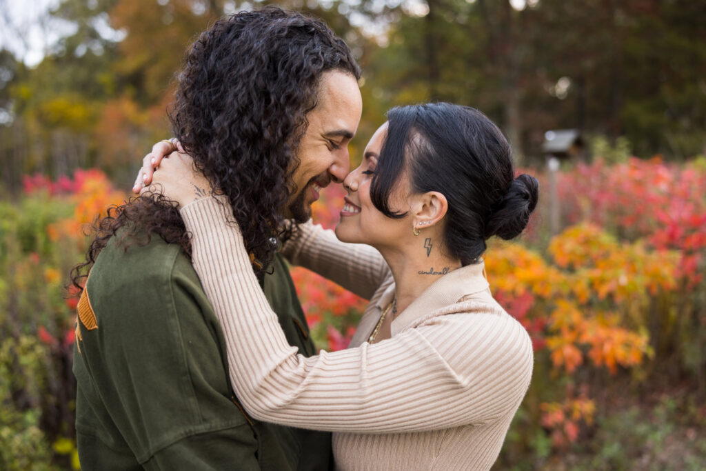 Engaged couple embracing and smiling forehead to forehead, surrounded by vibrant autumn foliage at Bethpage State Park.
