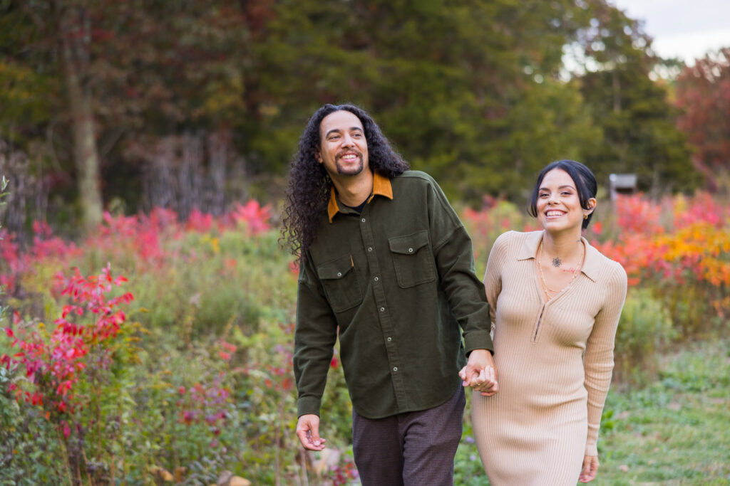 Playful couple holding hands and laughing while walking through colorful autumn meadows at Bethpage State Park.