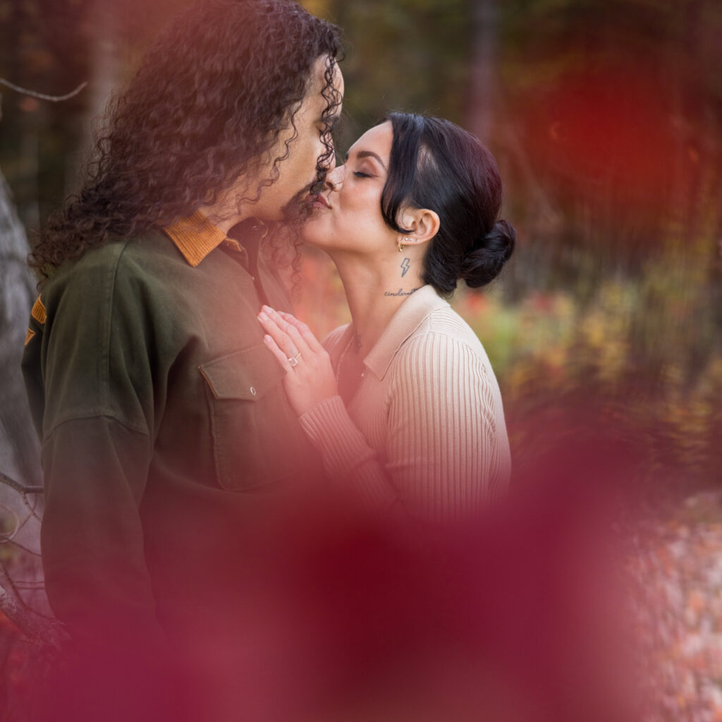 Engaged couple sharing a tender kiss framed by soft red autumn leaves at Bethpage State Park.