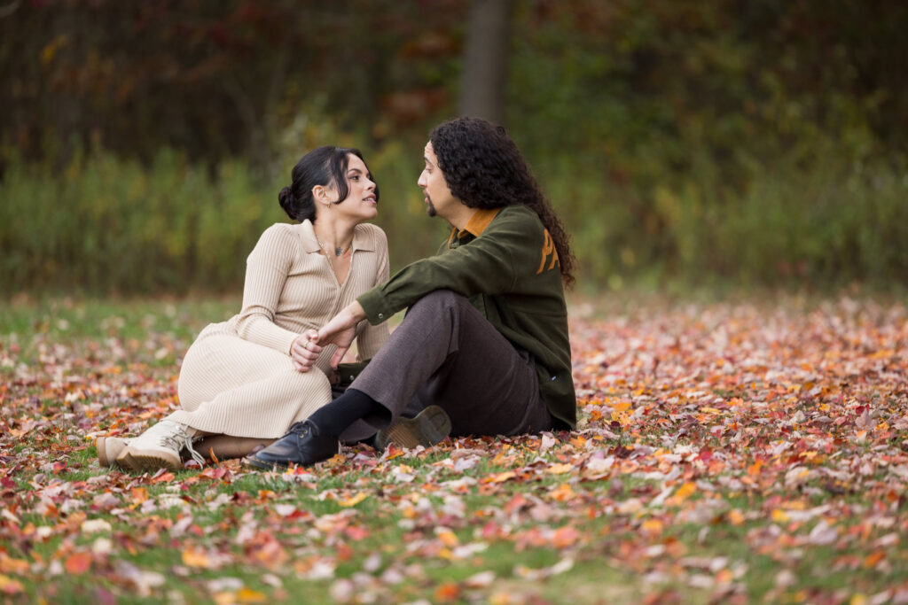 Engaged couple sitting close on a blanket of fallen autumn leaves, holding hands and smiling at Bethpage State Park.