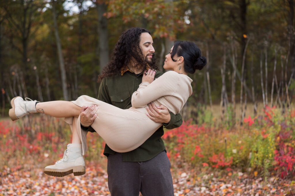 Fiancé lifting his bride-to-be in his arms as they share a loving gaze amidst colorful fall foliage at Bethpage State Park.