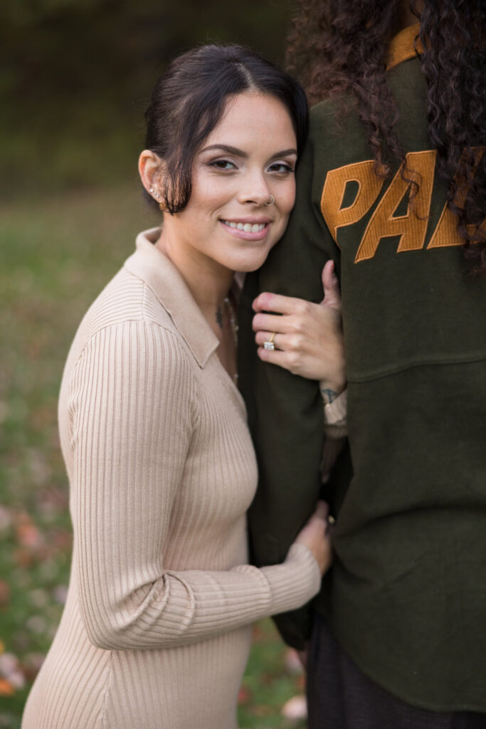 Bride-to-be smiling while holding her fiancé’s arm, showcasing her engagement ring during a cozy fall session at Bethpage State Park.