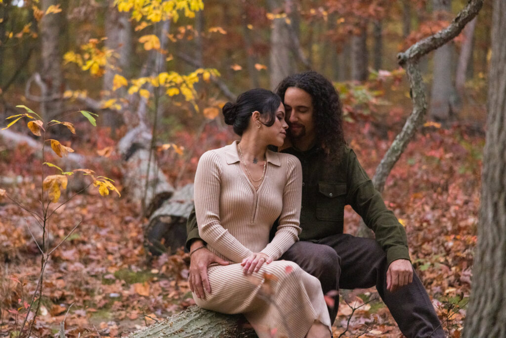 Engaged couple sitting closely on a fallen tree in a quiet forest clearing, surrounded by autumn leaves at Bethpage State Park.