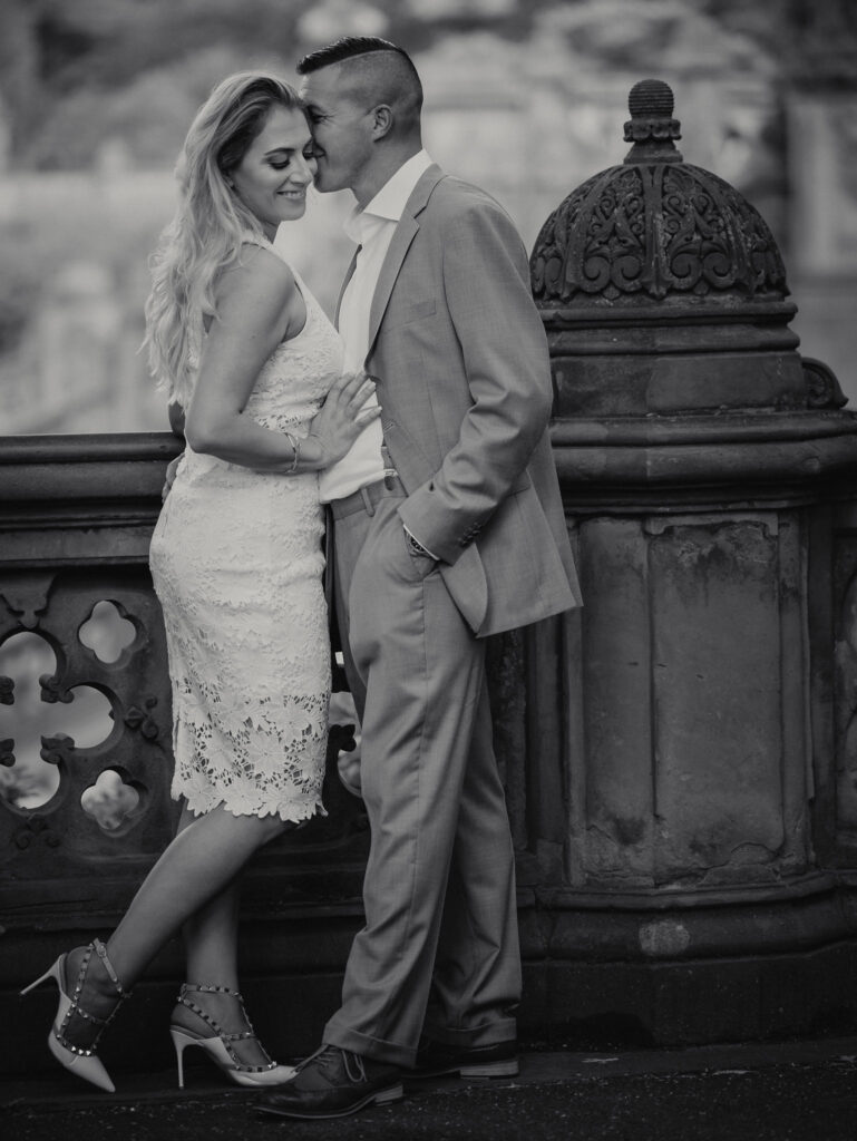 Black-and-white engagement photo of a couple embracing at Bethesda Terrace in Central Park, with the man in a tailored suit leaning in to kiss the woman in a lace dress and heels.
