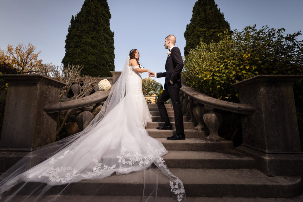 Bride and groom holding hands on an elegant stone staircase, with the bride’s long lace veil flowing behind her, captured to illustrate how to choose a wedding photographer with dramatic, cinematic wedding photography style in a garden estate setting.