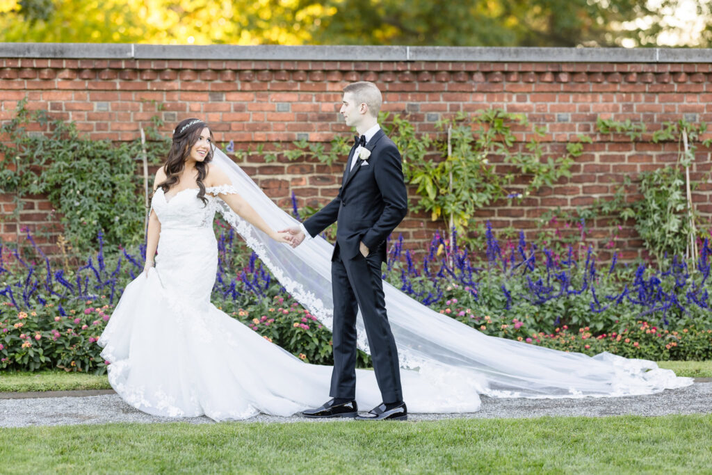 Bride and groom share a playful, romantic moment in a garden courtyard, holding hands in front of a brick wall as her long lace veil flows behind her, captured with documentary wedding photography.