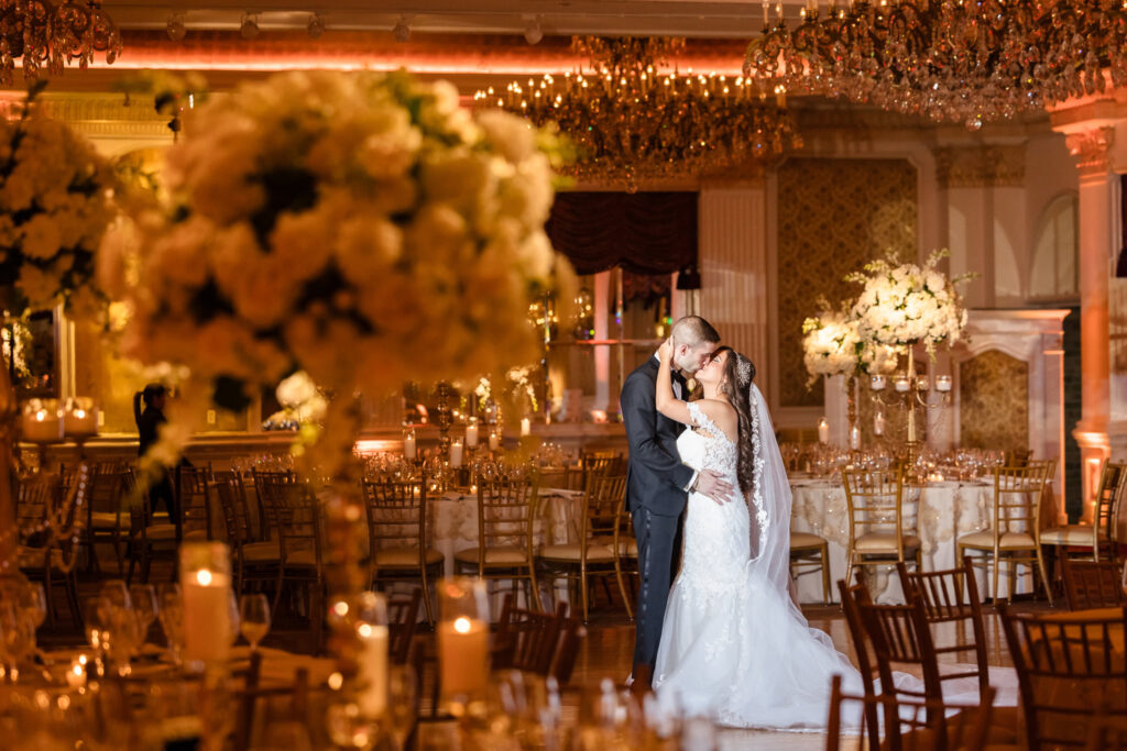 Bride and groom share a romantic kiss in an opulent ballroom, framed by candlelight and golden floral arrangements, with cinematic lighting highlighting their connection.