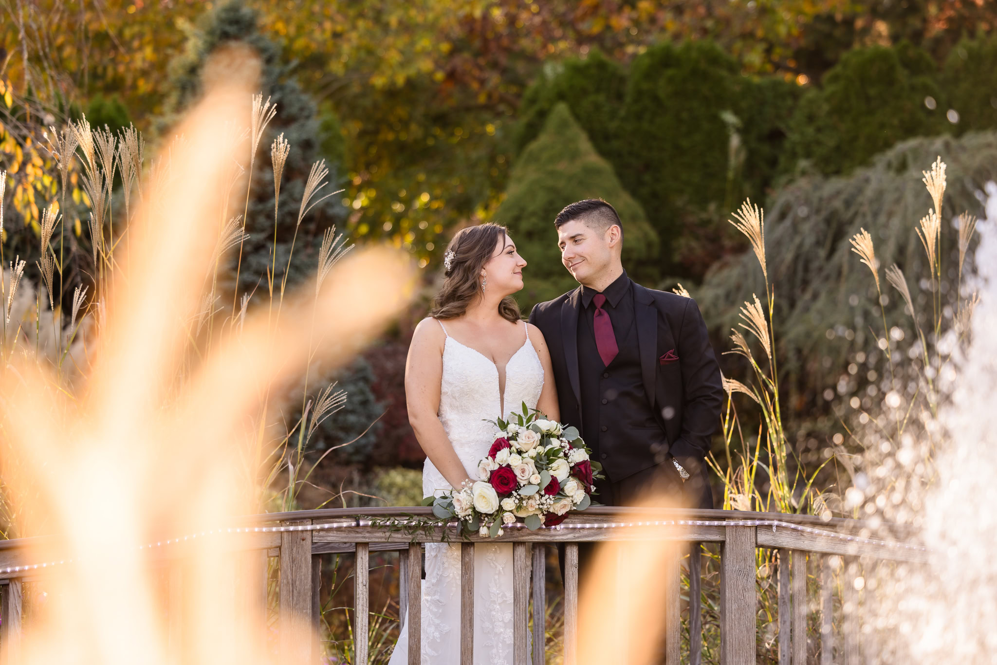 Bride and groom stand on a wooden bridge surrounded by fall foliage and ornamental grasses, sharing a quiet, joyful moment in a lush garden setting.