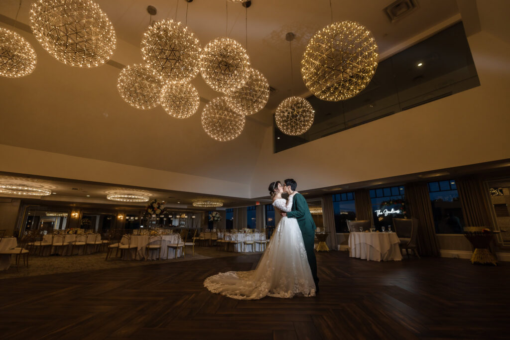 Bride and groom share their first dance beneath elegant orb chandeliers in a grand reception space, illuminated with cinematic lighting that highlights their joy and romance.