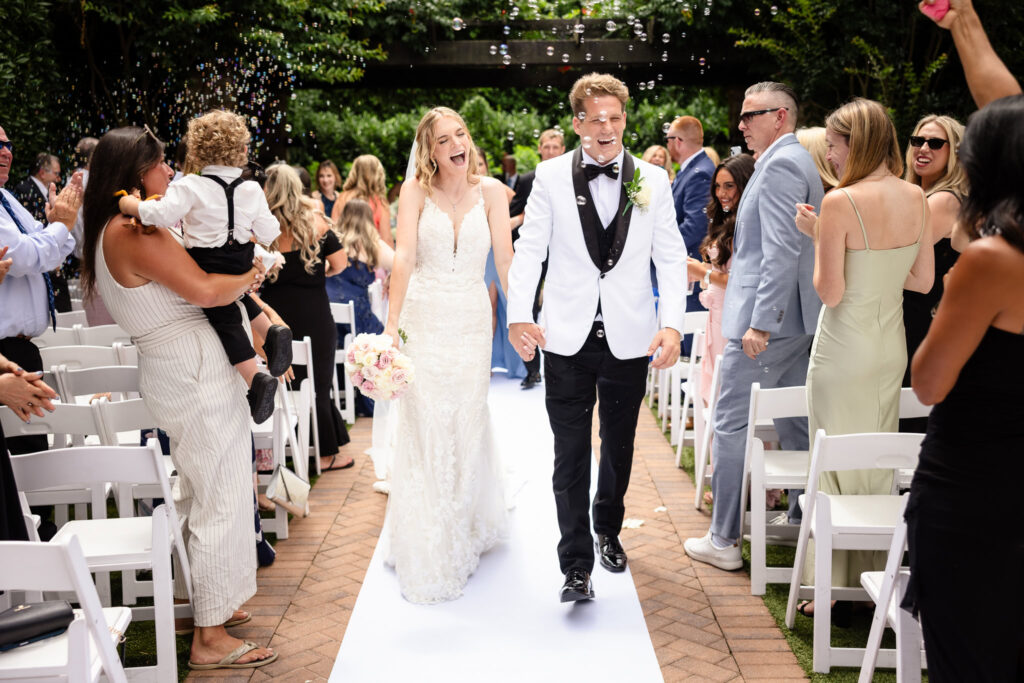 Bride and groom laughing joyfully as they walk down the aisle surrounded by cheering guests and bubbles.