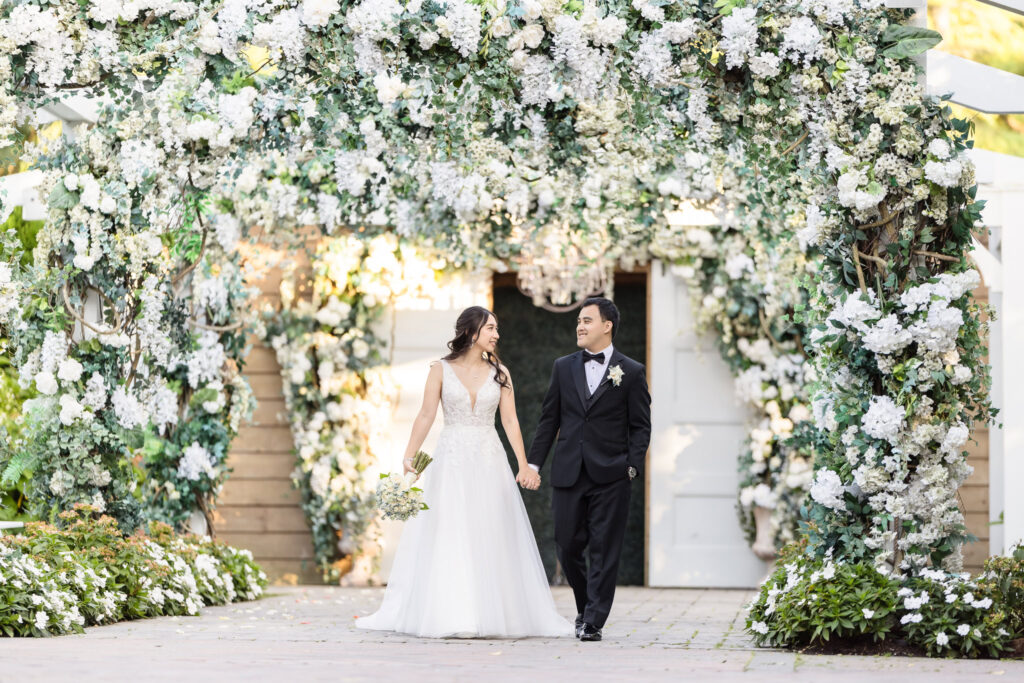 Newlyweds walking hand-in-hand under an archway of lush white florals, smiling at each other warmly.