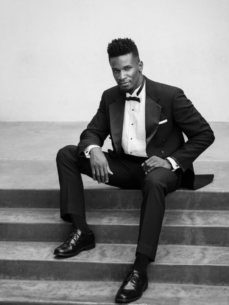 Stylish groom in a classic black tuxedo seated on stone steps, captured in a refined black and white portrait with editorial poise and timeless elegance.