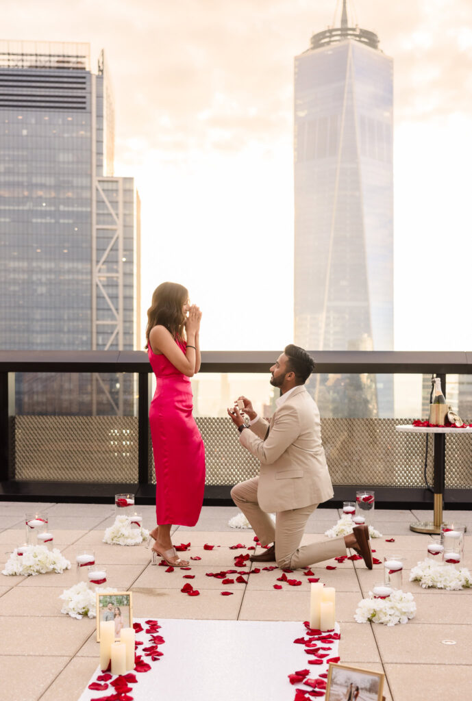 Romantic rooftop proposal in New York City at golden hour, with a groom-to-be kneeling amid rose petals as his emotional partner reacts with joy, captured with documentary wedding photography against the skyline.