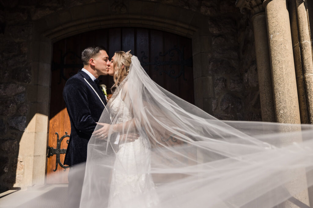 Bride and groom kissing outside a stone church entrance, with the bride’s long veil flowing dramatically in the sunlight during their wedding portraits.