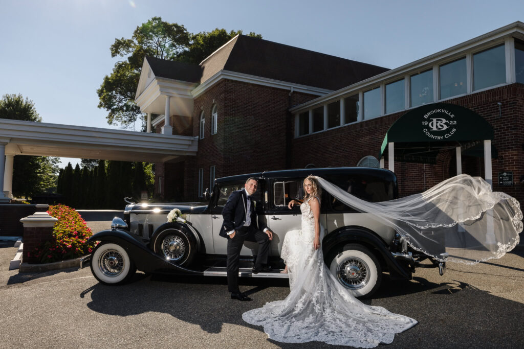 Bride and groom posing beside a vintage Rolls Royce at Brookville Country Club, with the bride’s lace veil flowing elegantly in the breeze, captured as a timeless editorial portrait with editorial wedding photography of luxury and legacy.